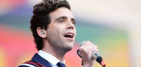 Singer Mika wearing a blue, white and red suit, white shirt and blue tie while performing on stage, with a grey microphone in his hand.