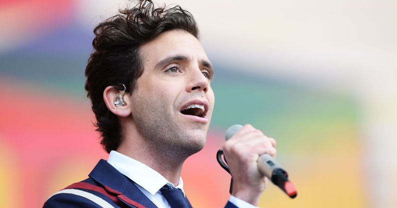 Singer Mika wearing a blue, white and red suit, white shirt and blue tie while performing on stage, with a grey microphone in his hand.
