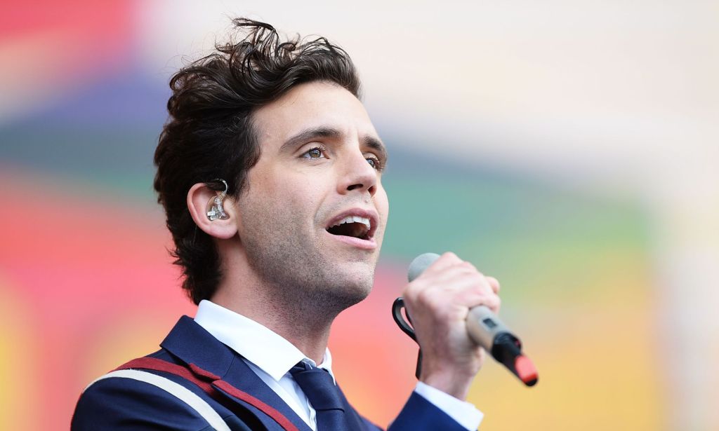 Singer Mika wearing a blue, white and red suit, white shirt and blue tie while performing on stage, with a grey microphone in his hand.