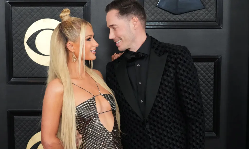 Paris Hilton, who is wearing a sparkly dress, smiles as she looks at her husband Carter Reum, who is wearing a black suit