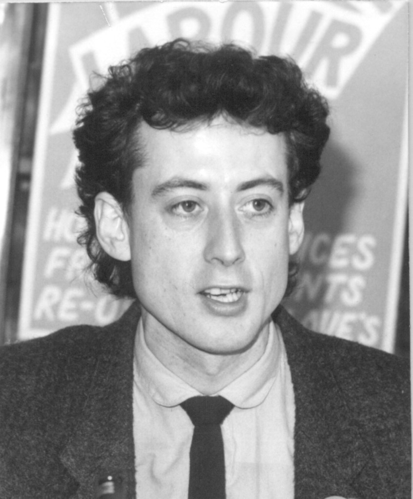 Peter Tatchell pictured at a press conference ahead of the Bermondsey by-election.  The black and white picture shows a young Tatchell wearing a suit as he addresses journalists.