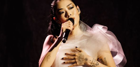 Rina Sawayama performs in a white dress while holding a black microphone.