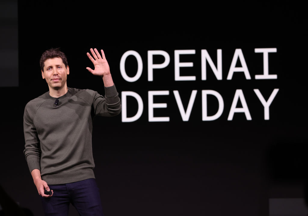 This is a photo of Sam Altman, the CEO of OpenAI. He is standing on a stage and is waving with his right hand. 
 
