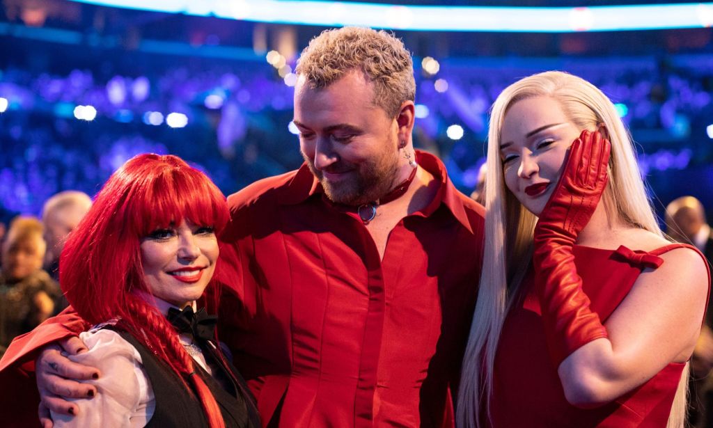 Shania Twain with red hair, Sam Smith with a red shirt, and Kim Petras with a red dress and red gloves, with their arms around each other at the 2023 Grammy awards.