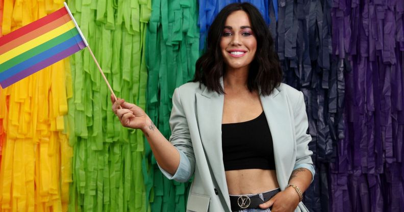 Sophie Cachia wearing a black top and grey blazer stands with one hand in her pocket and the other holding a pride flag.