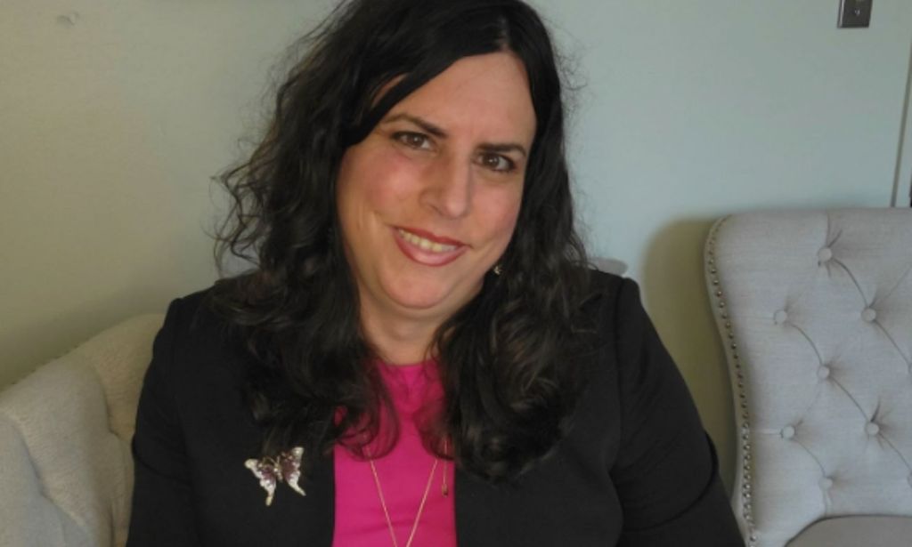 Photo of Dr Tiffany Najberg wearing a black suit jacket over a pink top as she sits on a chair