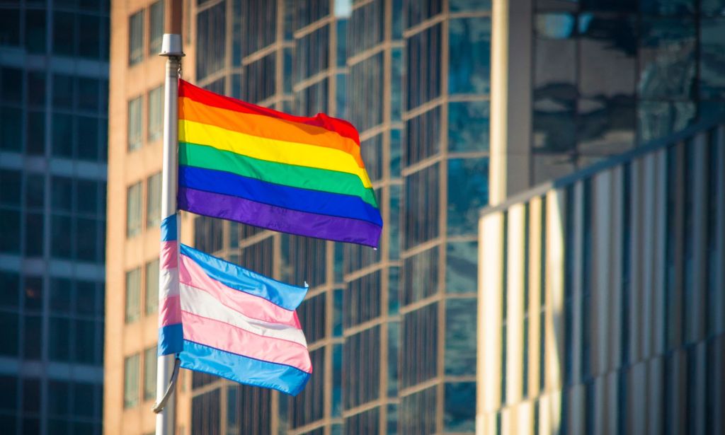 A pride flag is flying above a trans pride flag on a flagpole in an urban setting.