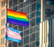 A pride flag is flying above a trans pride flag on a flagpole in an urban setting.