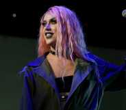 Screenshot of drag queen Adore Delano wearing a black bra top and grey overcoat with pink wig while she holds a microphone