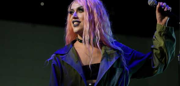 Screenshot of drag queen Adore Delano wearing a black bra top and grey overcoat with pink wig while she holds a microphone
