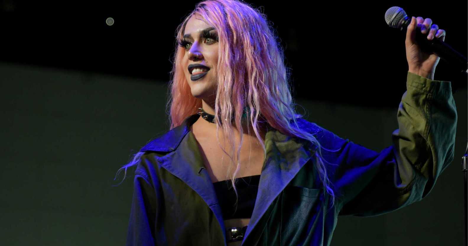 Screenshot of drag queen Adore Delano wearing a black bra top and grey overcoat with pink wig while she holds a microphone