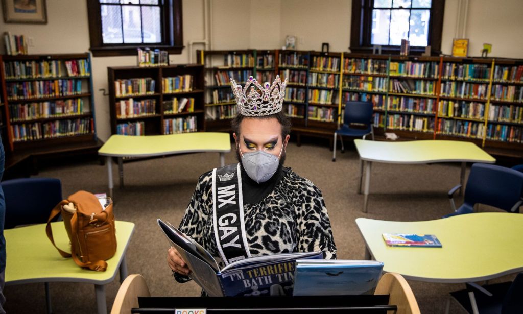 A drag queen reads from a book while standing in a library