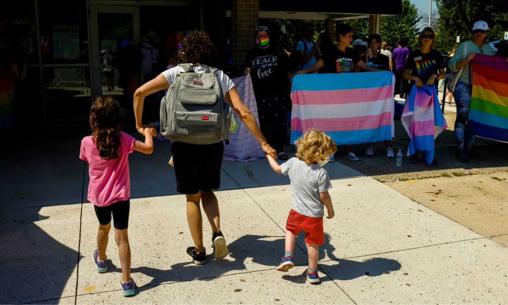 A parent holds the hands of two small children as they try to get into a drag event with people holding up a trans Pride flag to block out hateful protestors