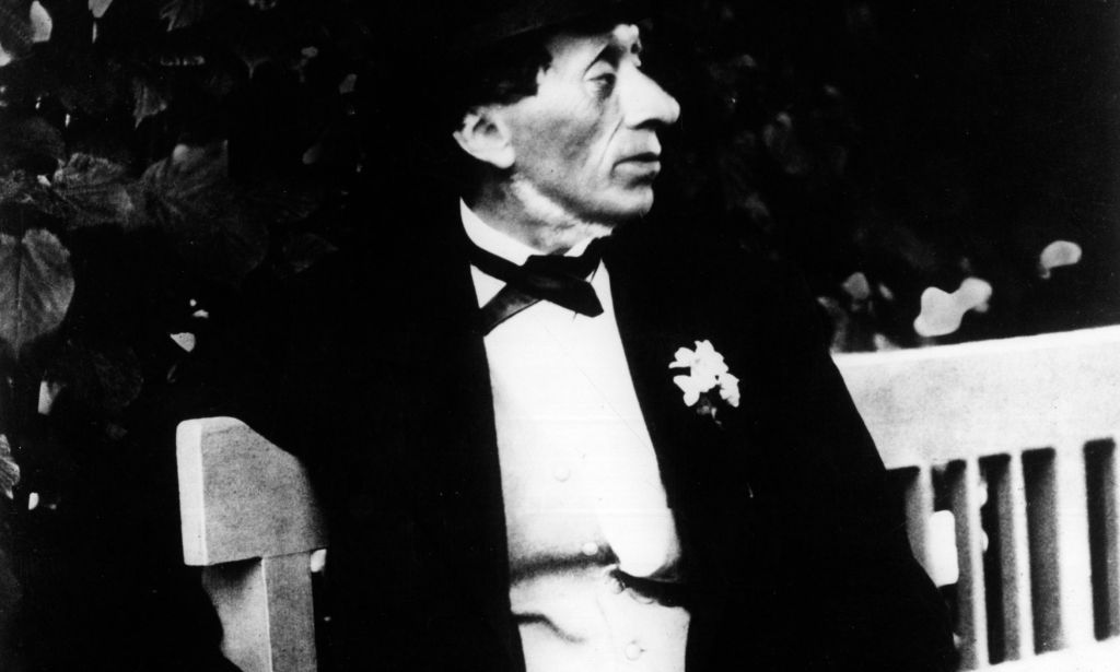 A black and white photograph of Danish author Hans Christian Andersen wearing a white shirt and dark clothing as he poses for the camera while sitting on a bench outside
