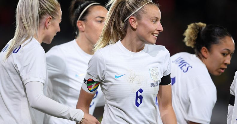 Lionesses captain Leah Williamson stands alongside her football teammates while wearing a white uniform and rainbow OneLove armband