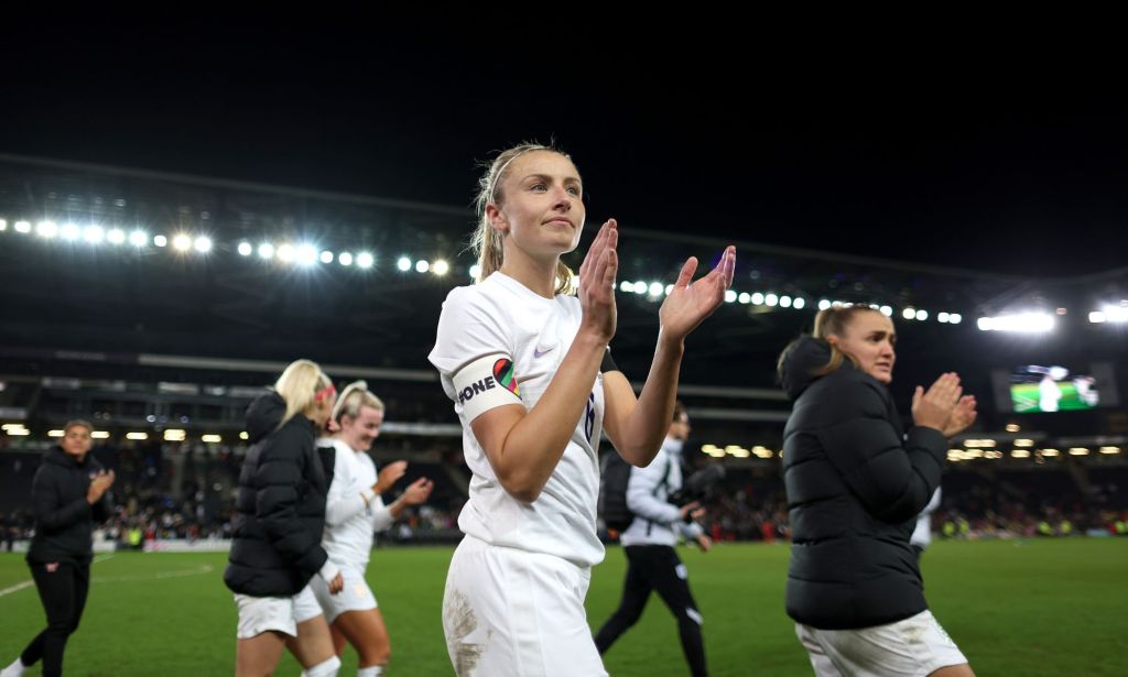 Lionesses captain Leah Williamson claps as she walks off the football field while wearing a white uniform and the rainbow OneLove armband
