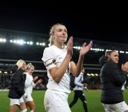 Lionesses captain Leah Williamson claps as she walks off the football field while wearing a white uniform and the rainbow OneLove armband