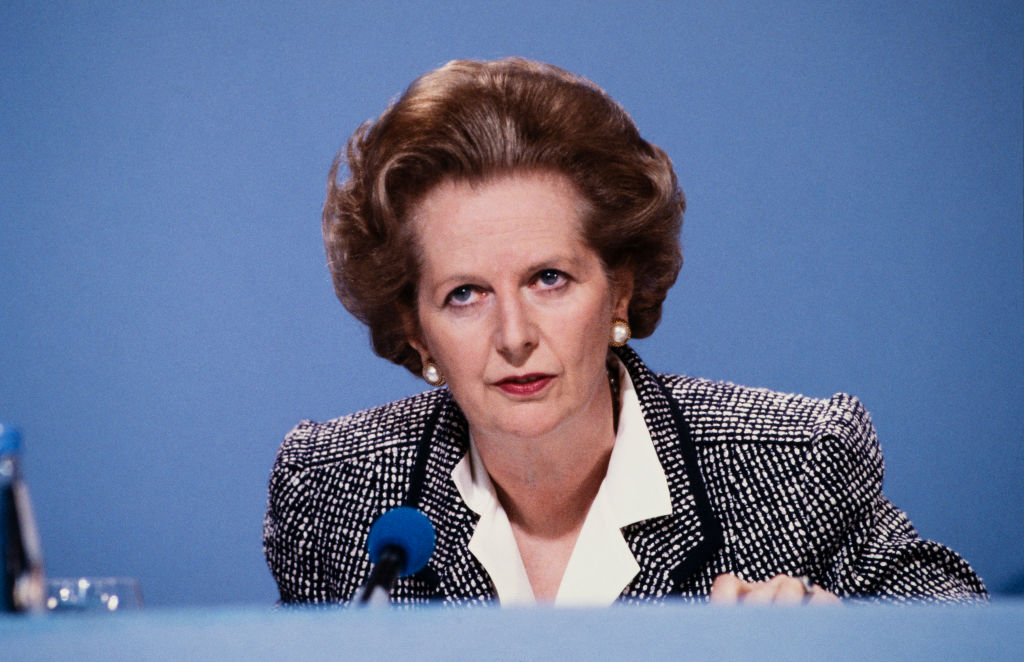Margaret Thatcher in a grey suit and white shirt, sitting in front of a blue backdrop