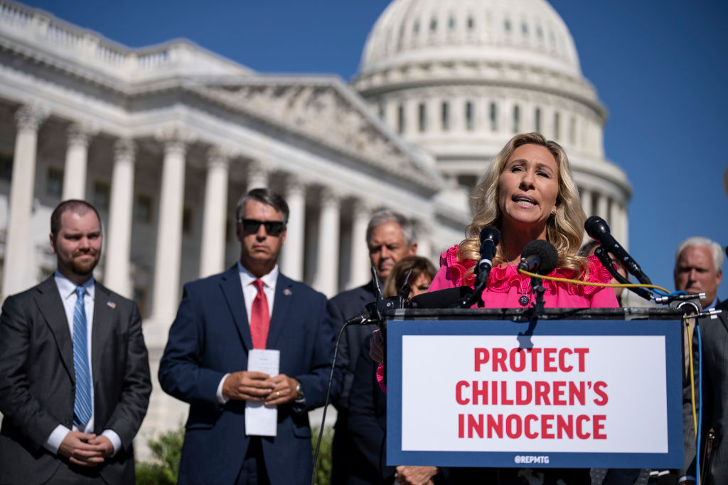 Marjorie Taylor Greene is one of the most vocal anti-trans politicians in the United States. She is pictured here outdoors at Congress taking part in a demonstration against trans rights. She is standing at a podium with a sign that reads "protect children's innocence".