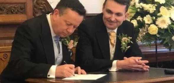 Paul and Alain in suits, sat at a dark wood table, signing their marriage certificate