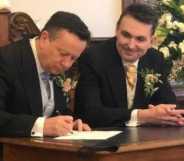Paul and Alain in suits, sat at a dark wood table, signing their marriage certificate