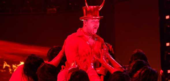A screenshot from the 2023 Grammys ceremony shows Sam Smith performing "Unholy" on stage wearing a hat showing devil horns as a red spotlight casts a strong red light over him
