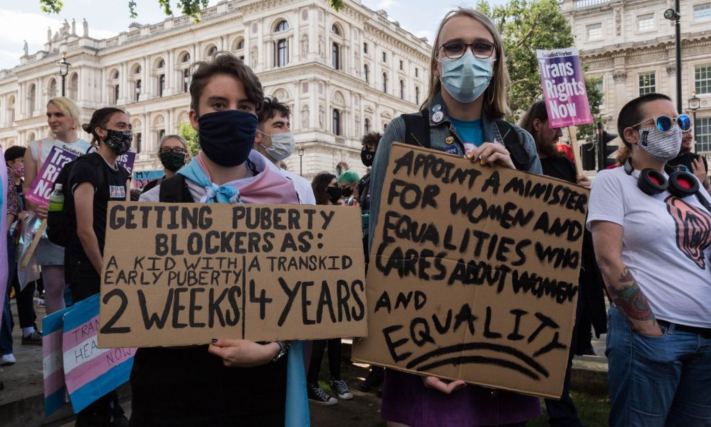 Two people are pictured holding up signs in support of the trans community at a protest. One person's sign reading criticises the extensive wait times trans youth face accessing GIDS services through the NHS versus the shorter times cisgender people must wait for similar treatment