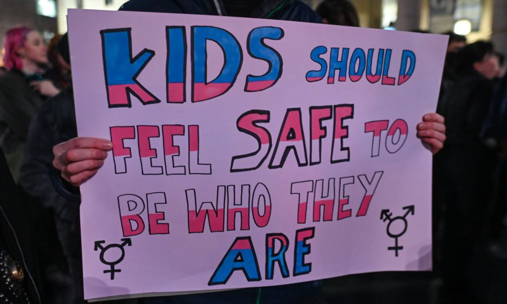 A person holds a sign reading "kids should feel safe to be who they are" in the colour of the trans pride flag