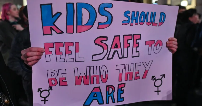 A person holds a sign reading "kids should feel safe to be who they are" in the colour of the trans pride flag