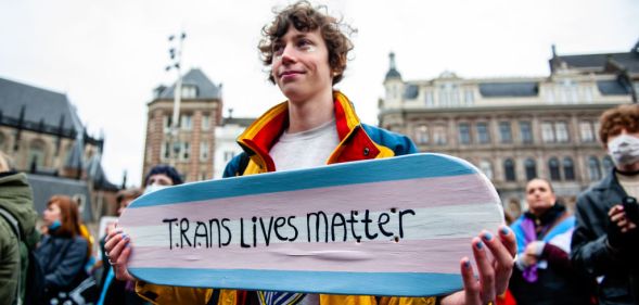 A person is holding a skateboard with the colors of the transgender flag and with a message in support of trans people.