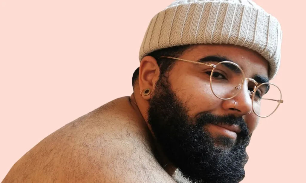 Bappie Kortram, a Black, fat, trans model and designer based in the Netherlands smiles as he wears a cream-coloured beanie hat in a shirtless photo