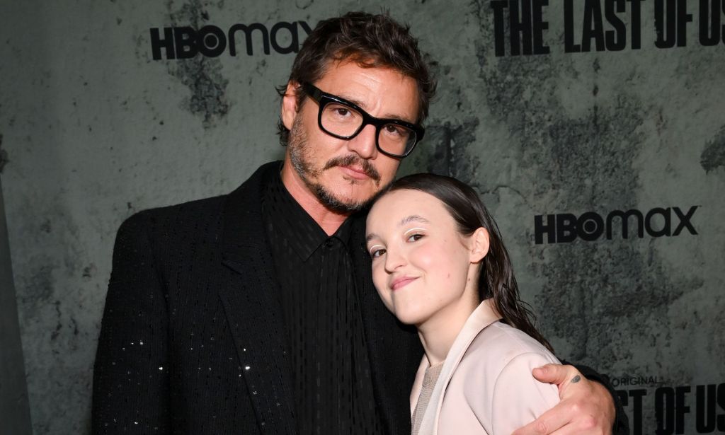 Pedro Pascal in a black suit and black glasses hugging Bella Ramsey, who is smiling and wearing a light pink suit.