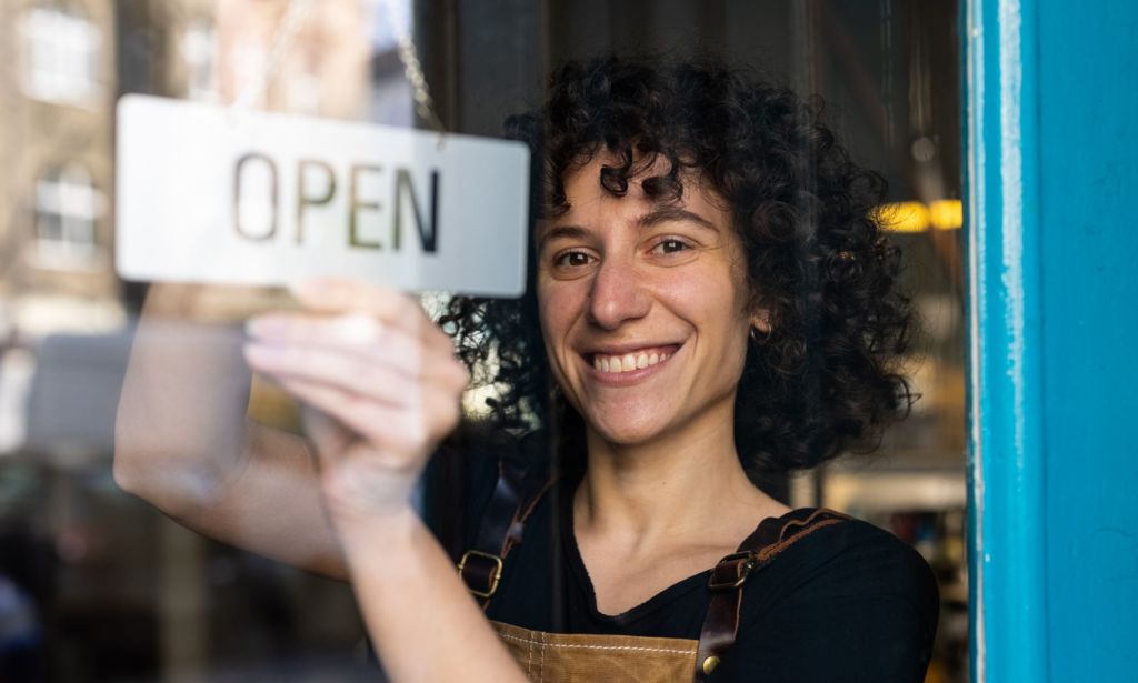 A woman with curly hair is turning over an "open" sign behind a glass door.
