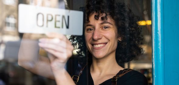 A woman with curly hair is turning over an "open" sign behind a glass door.