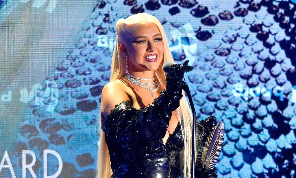 Christina Aguilera in a blue dress holding a microphone and her GLAAD Award stood against a blue sequinned background.