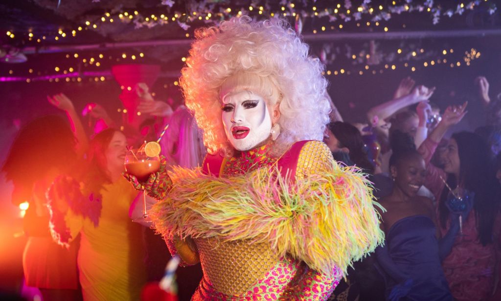 Danny Beard in a fluffy yellow and pink top and blonde wig, holding a cocktail.