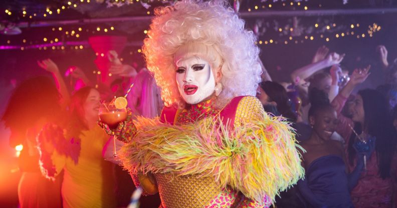 Danny Beard in a fluffy yellow and pink top and blonde wig, holding a cocktail.