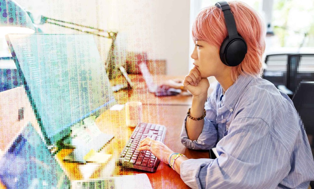 An asian woman with pink hair is wearing headphones and looking at a computer monitor.