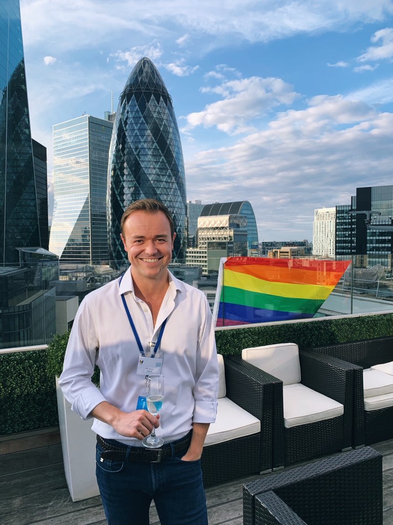 Ed johnson is on a rooftop in central london, wearing a white shirt and jeans. He is holding a glass and behind him is the pride flag.