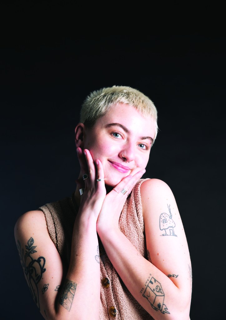 Content creator Ella Willis is posing for the camera on a black background. They are smiling for the camera and have their arms in their hands.