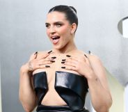 Mae Muller wears a black dress at the MTV 2022 VMAs, looking off camera, hands on her shoulders, and with her mouth open.
