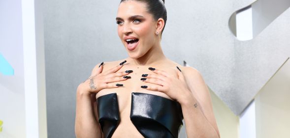 Mae Muller wears a black dress at the MTV 2022 VMAs, looking off camera, hands on her shoulders, and with her mouth open.