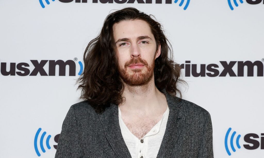Hozier wearing a white shirt and grey cardigan against a white and black background.