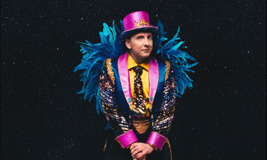 Joe Lycett in a vibrant pink, yellow and blue suit with blue feathers on the arms and a pink top hat.