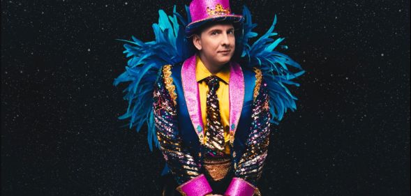 Joe Lycett in a vibrant pink, yellow and blue suit with blue feathers on the arms and a pink top hat.