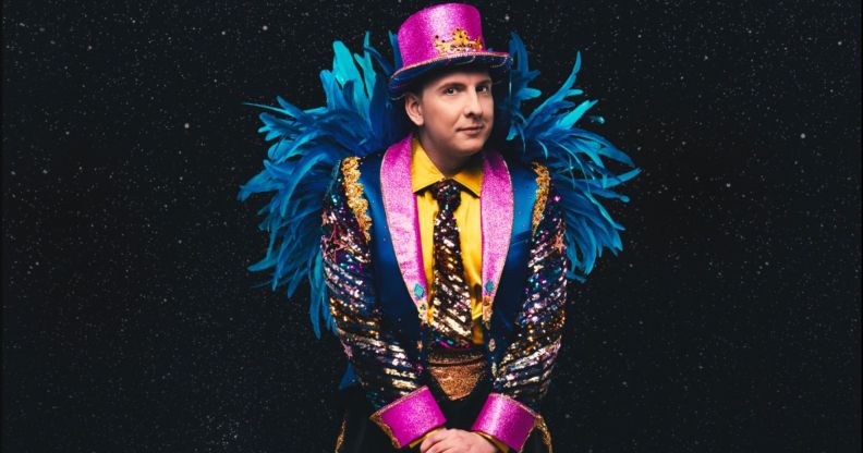 Joe Lycett in a vibrant pink, yellow and blue suit with blue feathers on the arms and a pink top hat.