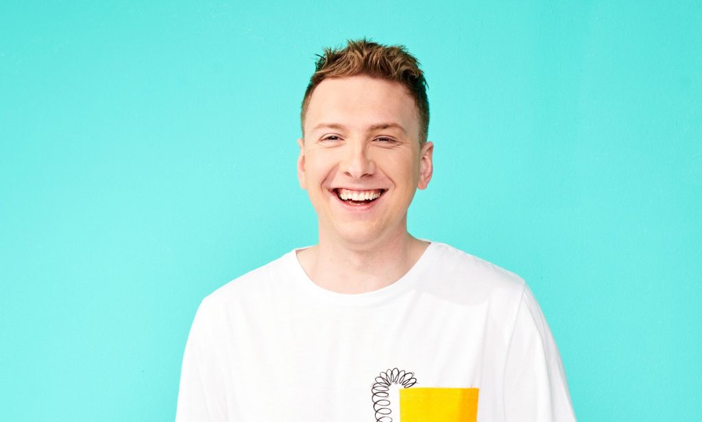 Joe Lycett wears a white and yellow t-shirt while stood against a light blue background and laughing.