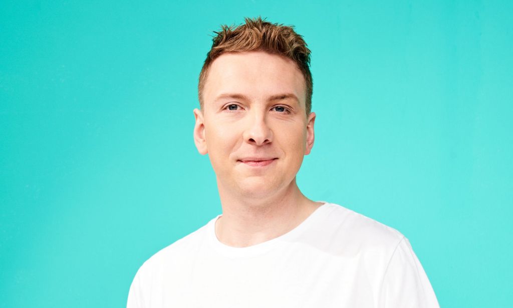 Joe Lycett wearing a white t-shirt and smiling at the camera against a teal background.