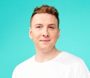 Joe Lycett wearing a white t-shirt and smiling at the camera against a teal background.