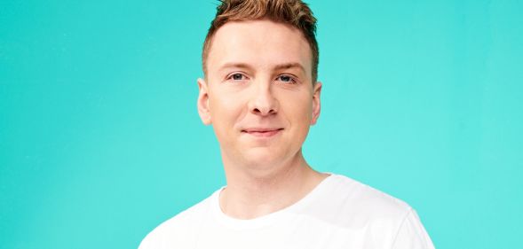 Joe Lycett wearing a white t-shirt and smiling at the camera against a teal background.
